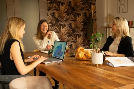 Visuels d'un conseiller immobilier ou d'une équipe en train de présenter des plans ou des documents à un couple de clients dans un intérieur de bureau ou d'agence immobilière. Maquettes architecturales, plans sur table, documents contractuels visibles, ambiance professionnelle et bienveillante. Le focus doit être sur l'interaction conseil et l'accompagnement personnalisé, sans texte ou logo prominent. Lumière naturelle, composition frontale ou de trois-quarts montrant l'échange d'informations entre professionnels et clients.